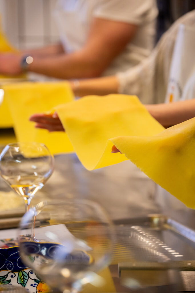 Hands holding sheets of fresh yellow pasta dough, a glass of white wine on the counter