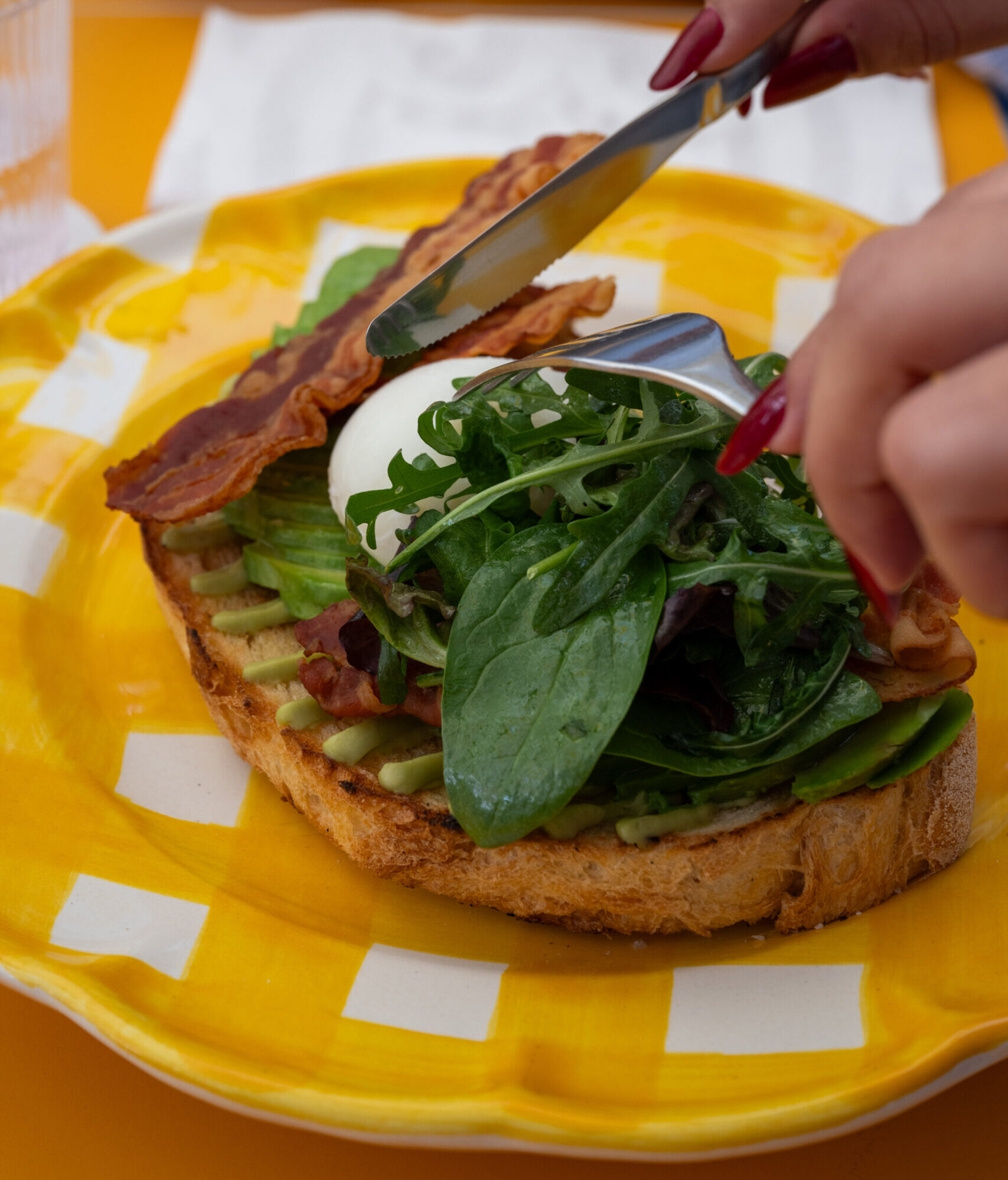 Avocado toast with bacon, poached egg and rocket on a yellow checked plate, cut with knife and fork